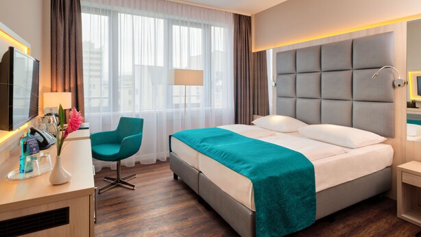 Una habitación de hotel con cama, escritorio, silla, lámpara y ventana con cortinas.