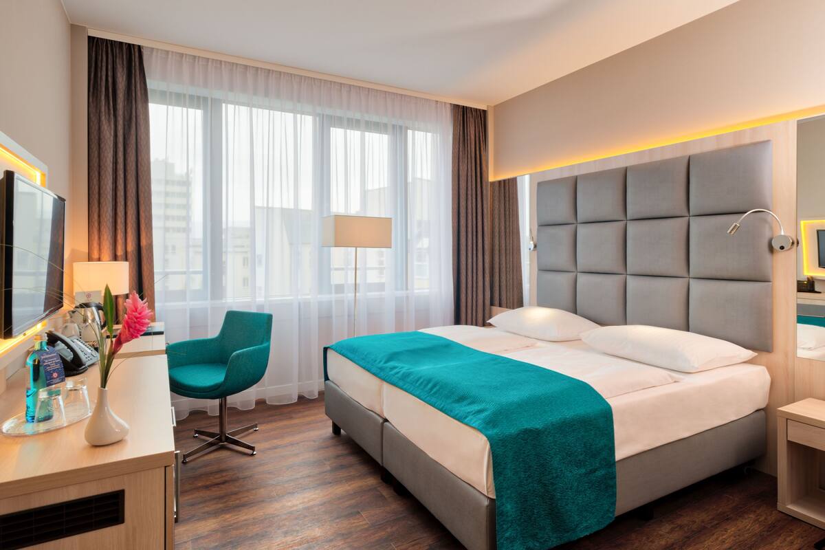 Quarto de hotel com uma cama, uma escrivaninha com cadeira, uma luminária e uma janela com cortinas.