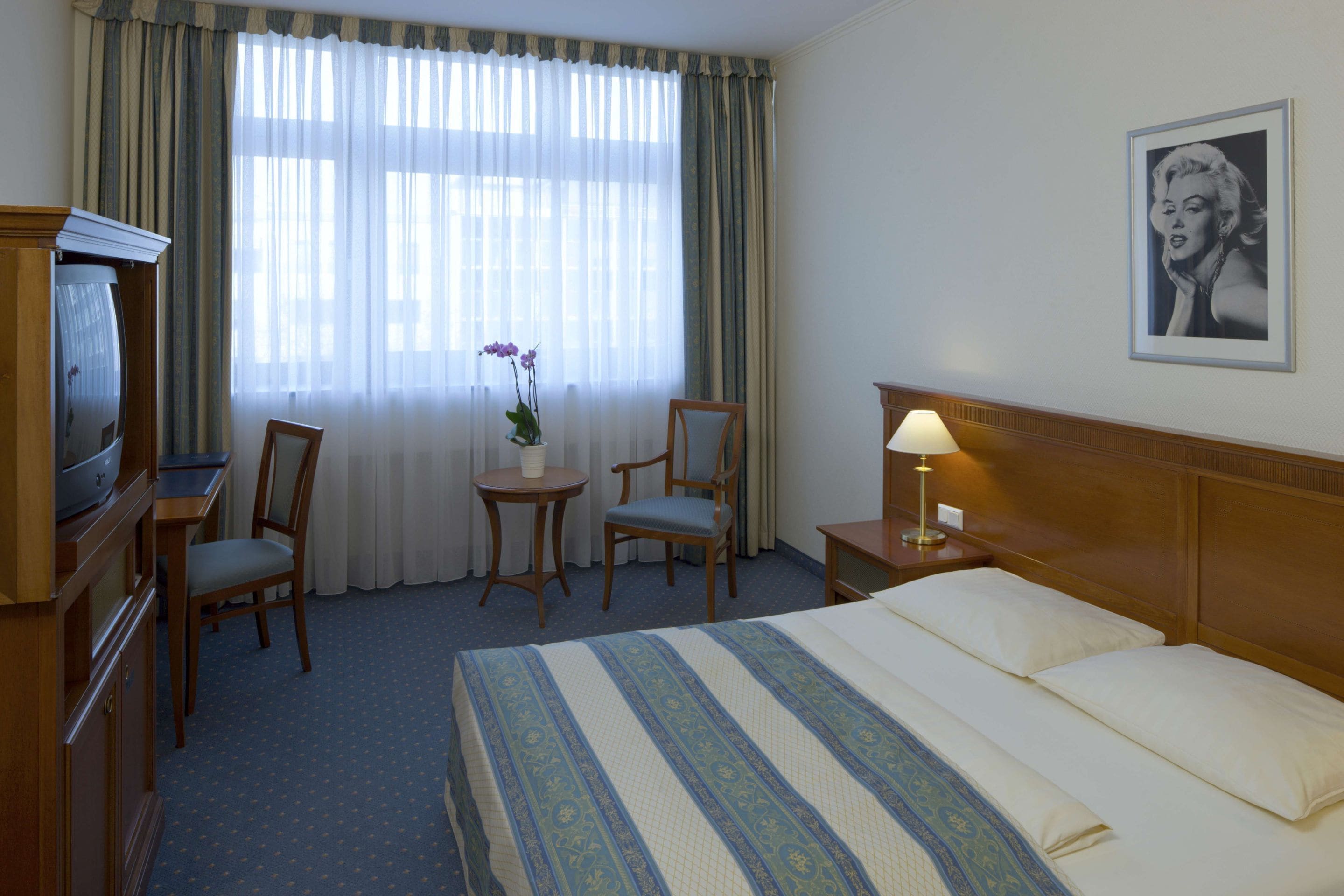 Habitación de hotel con cama, escritorio con televisor, dos sillones, mesita con un florero y lámpara en una mesita de noche.