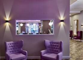 A reception area with a large mirror, two purple armchairs, and wall-mounted lights.