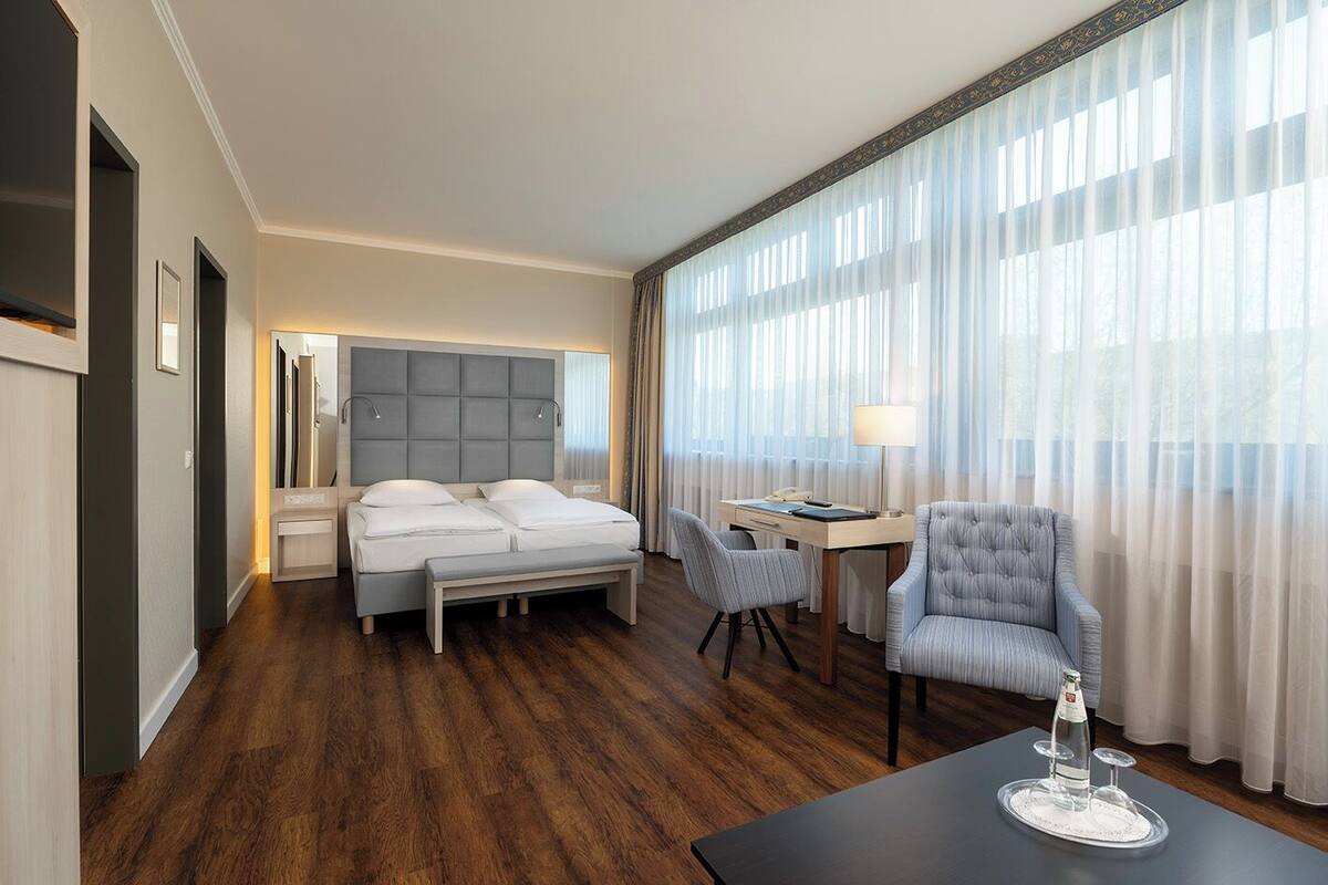 Quarto de hotel com cama, escrivaninha, cadeira e mesa com garrafa e copos.