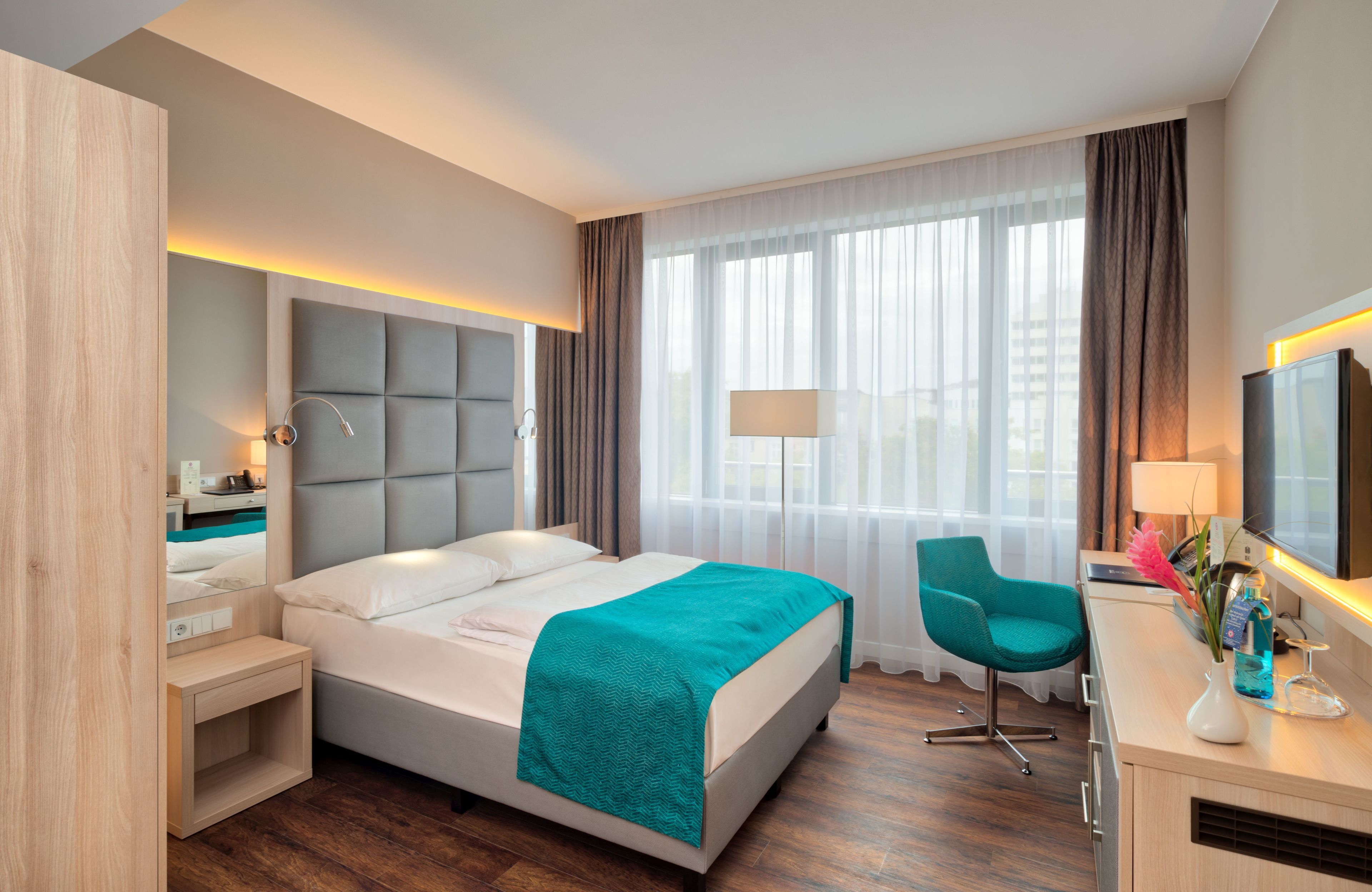 Una habitación de hotel moderna con cama, mesita de noche, silla y un ventanal con cortinas.