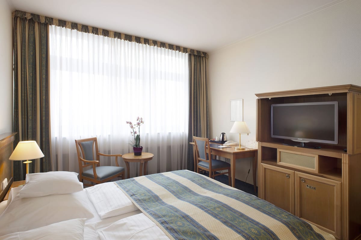 Quarto de hotel com cama, escrivaninha, cadeira, televisão e pequena mesa com abajur.