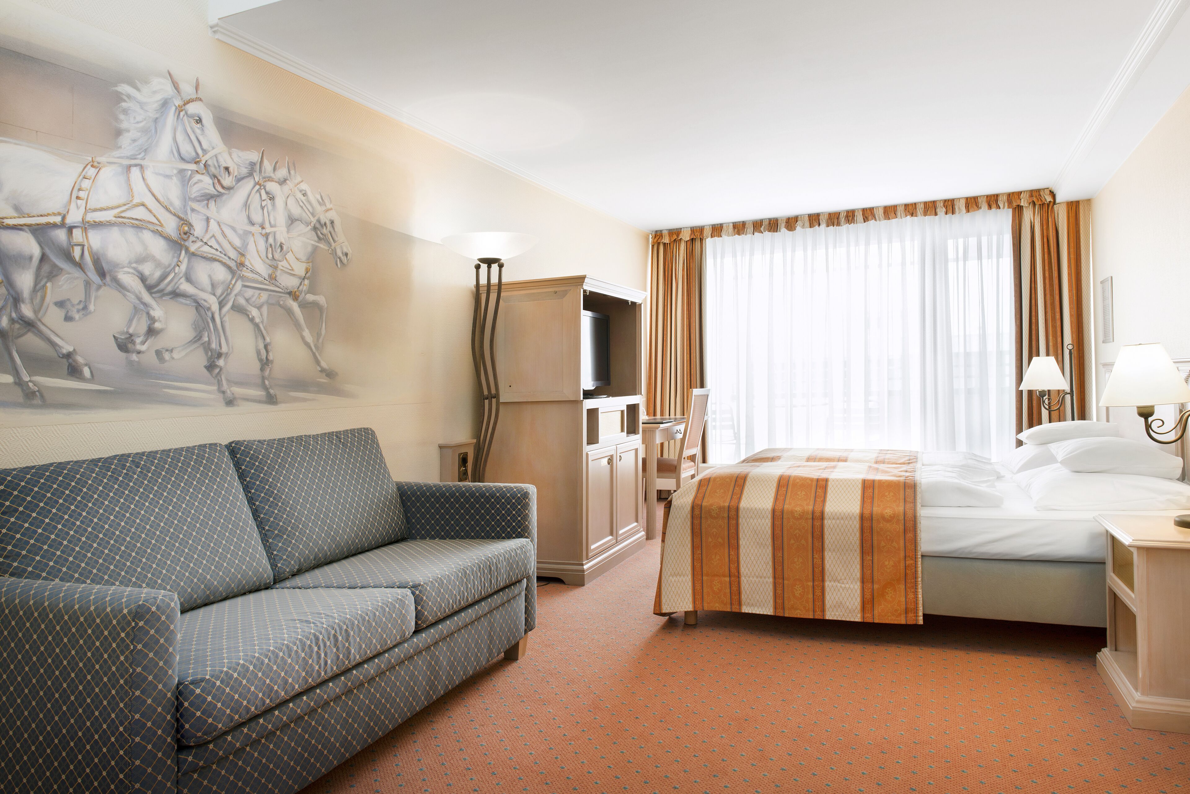 A hotel room with a bed, a sofa, a television, and a wall-mounted artwork of horses.