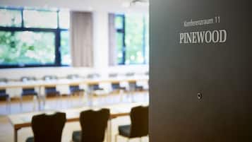 A conference room with tables and chairs, a large window with a view of greenery, and a sign indicating 'Konferenzraum 11 PINEWOOD'.