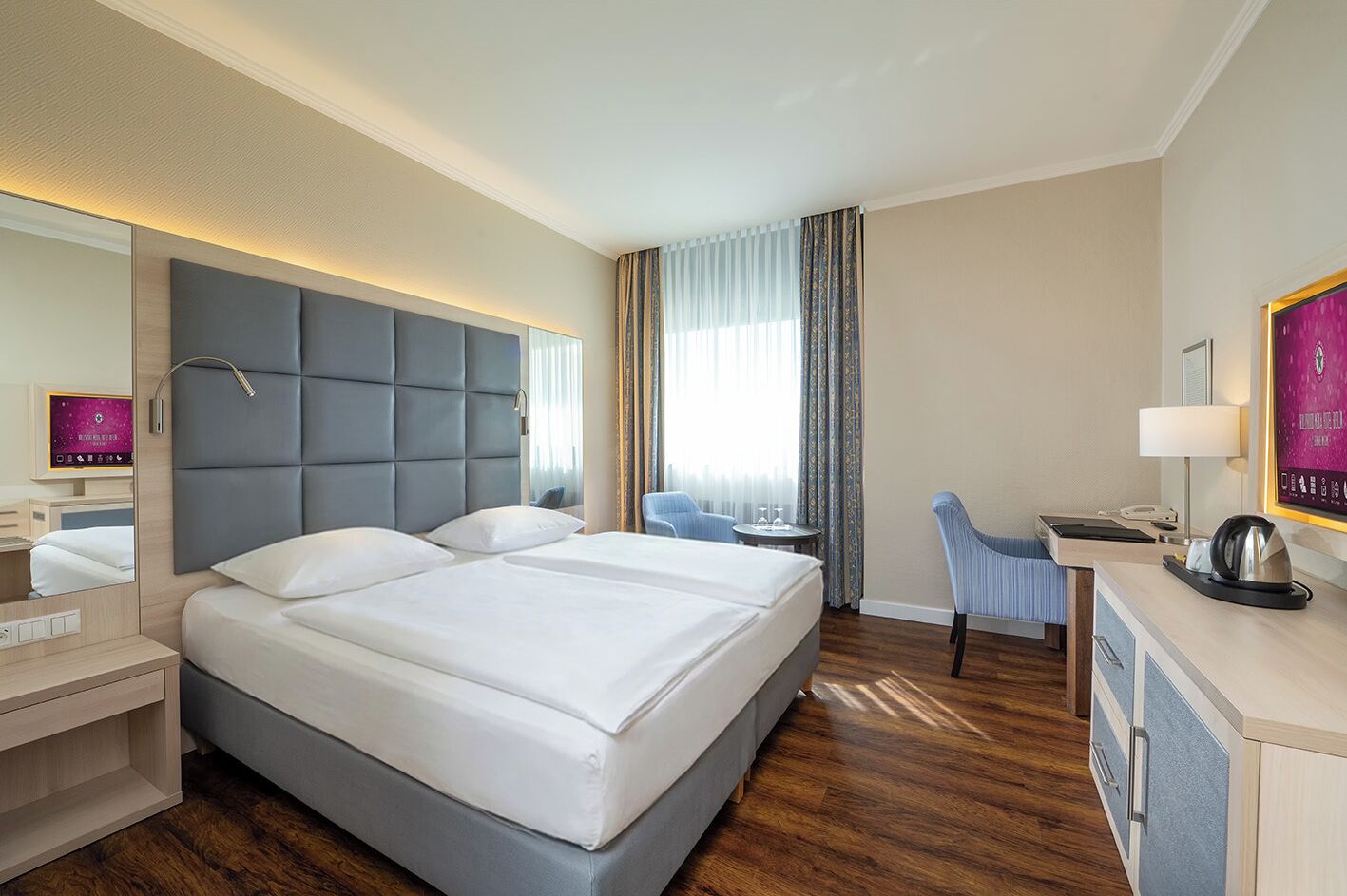 A hotel room with a large bed, a desk with a chair, a mirror, and a window with curtains.
