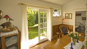 A dining room with a wooden table, chairs, and a vase of flowers. There is a window with a view of greenery outside. - The Duck House (Burnie)