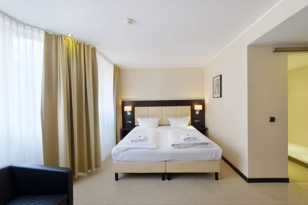 A hotel room with a large bed, two bedside tables with lamps, a chair, and a view of the outside through sheer curtains.