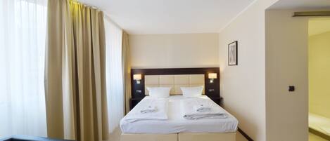 A hotel room with a large bed, two bedside tables with lamps, a chair, and a view of the outside through sheer curtains.