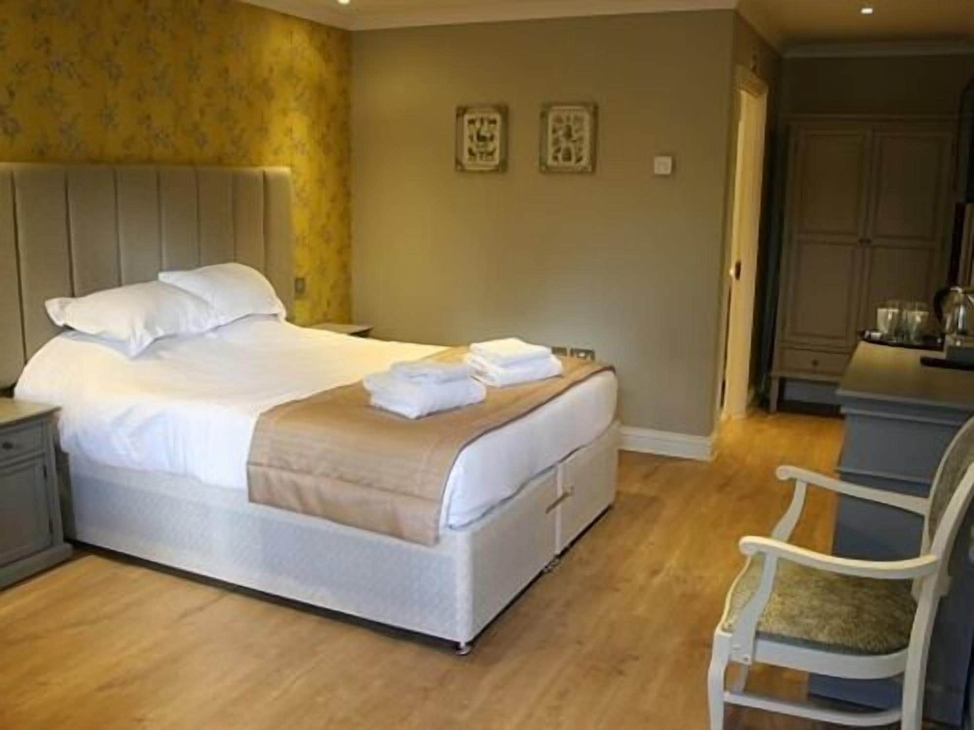 A neatly made bed with white linens and a beige throw blanket, a white chair with a cushion, and a wooden floor.