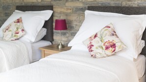 Dos camas con ropa blanca y almohadas florales, una lámpara roja y una pared de piedra al fondo.