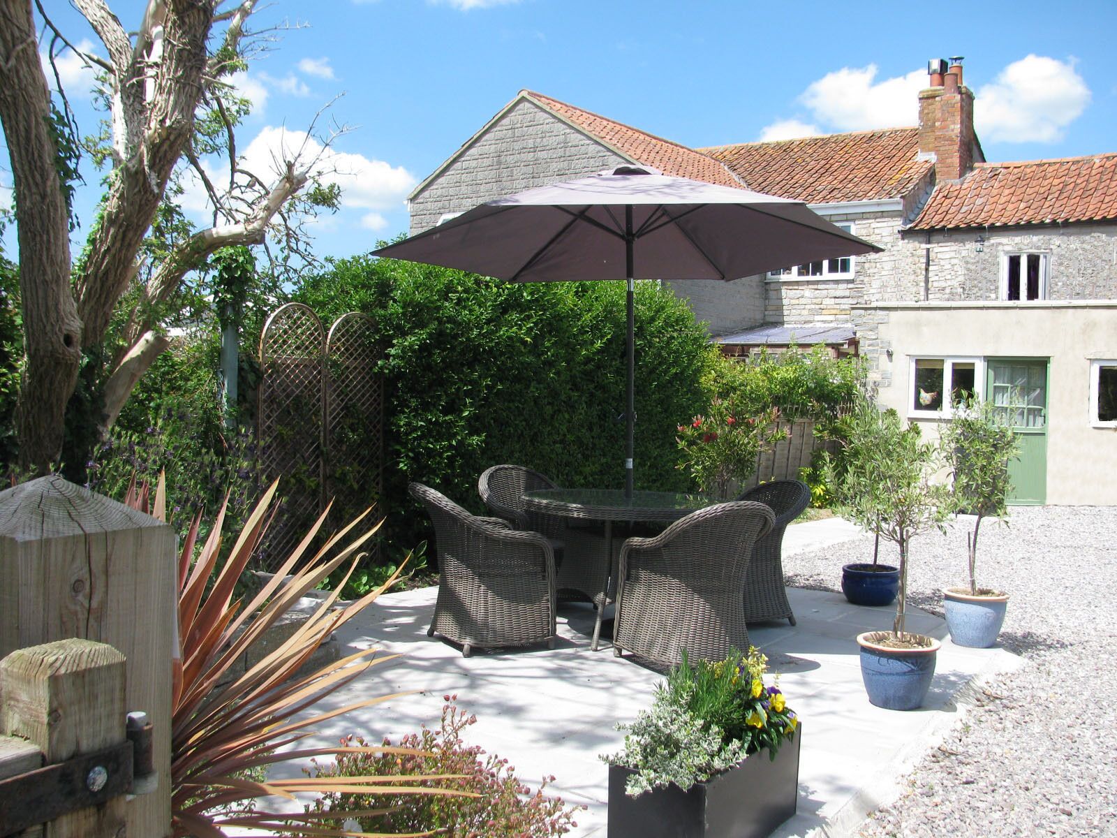 An outdoor patio with wicker furniture, a table, and an umbrella. There are potted plants and a stone wall in the background.