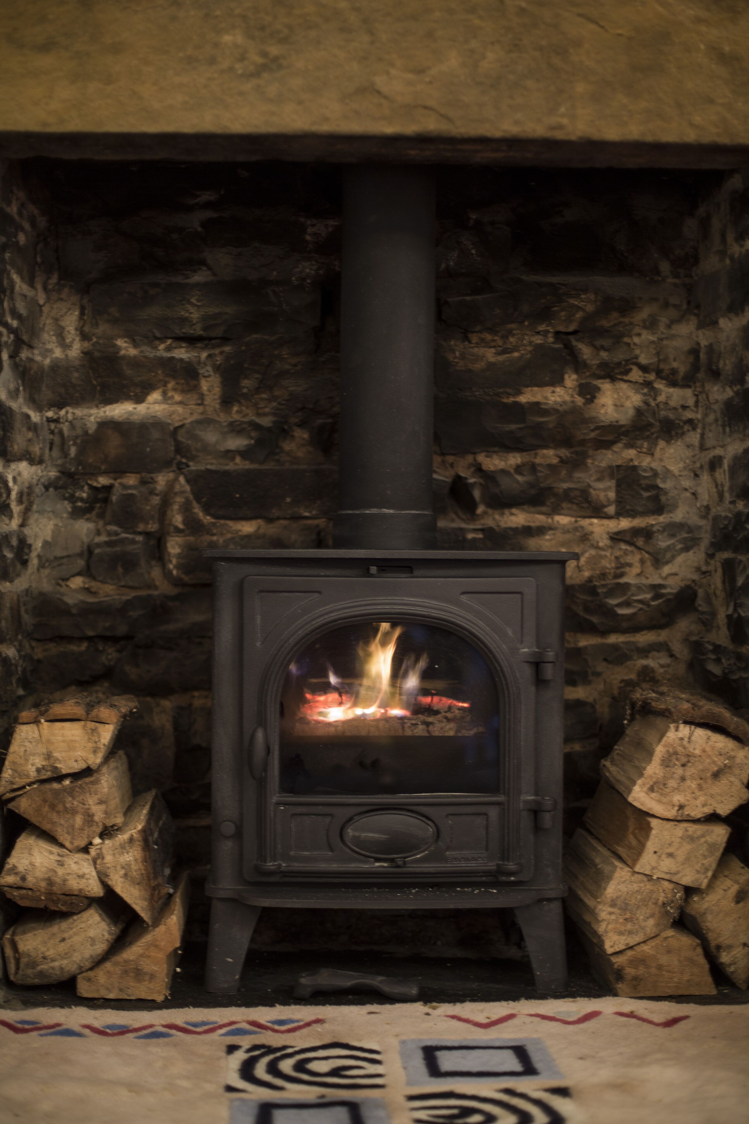 A wood-burning stove in a stone-walled room with logs stacked beside it.