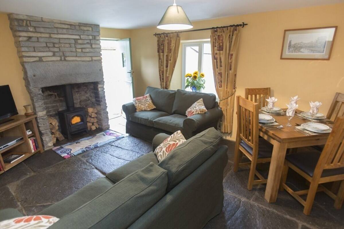 A cozy living room with a stone fireplace, a green sofa, a dining table set for four, and a bookshelf.
