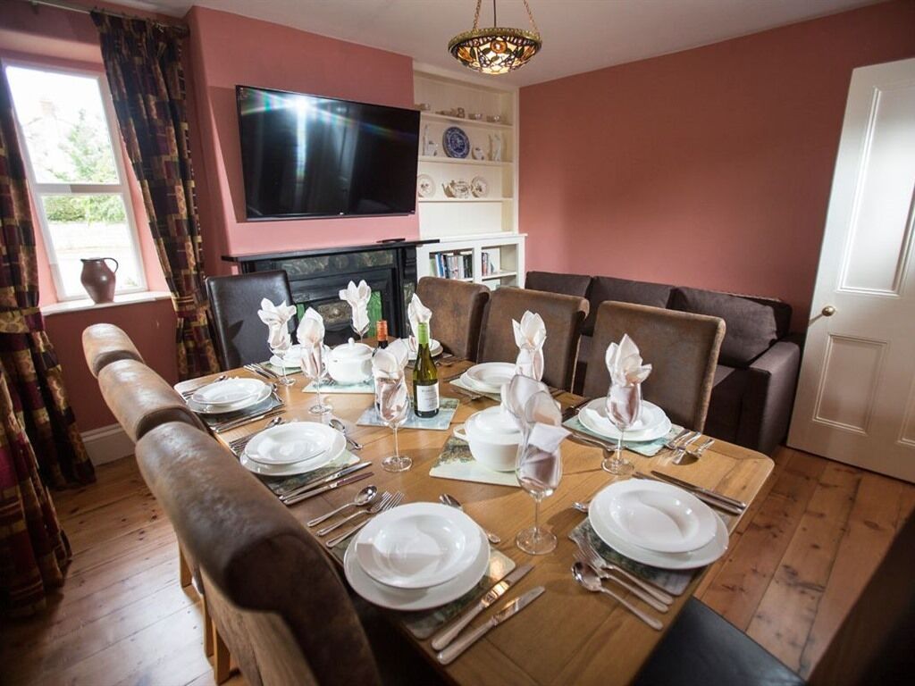 A dining room with a wooden table set for six, a television above the fireplace, and a sofa in the background.