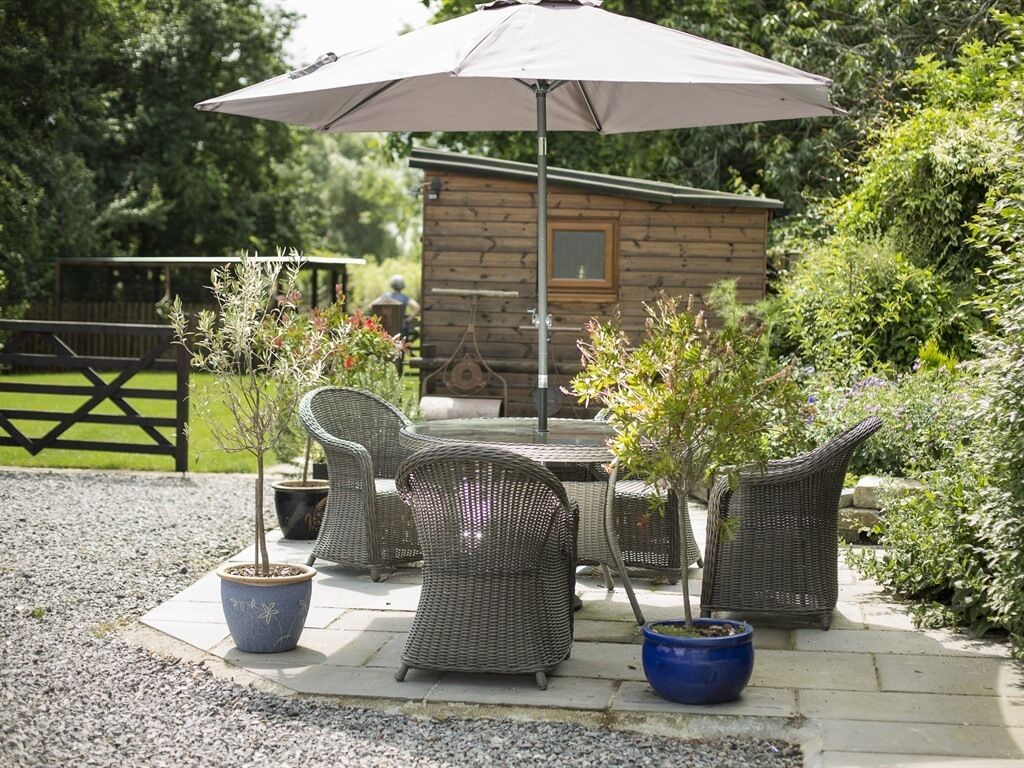 Un espace extérieur aménagé avec des chaises en osier et une table placée sous un grand parasol, entouré de plantes en pot et, au fond, d’une cabane en bois.