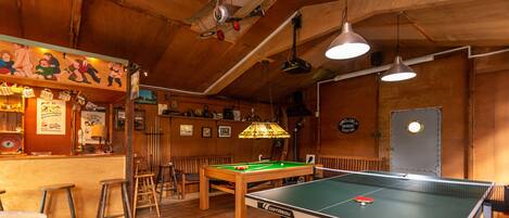 A billiards table with a pool cue, a bar area with stools, and a wooden ceiling with exposed beams.