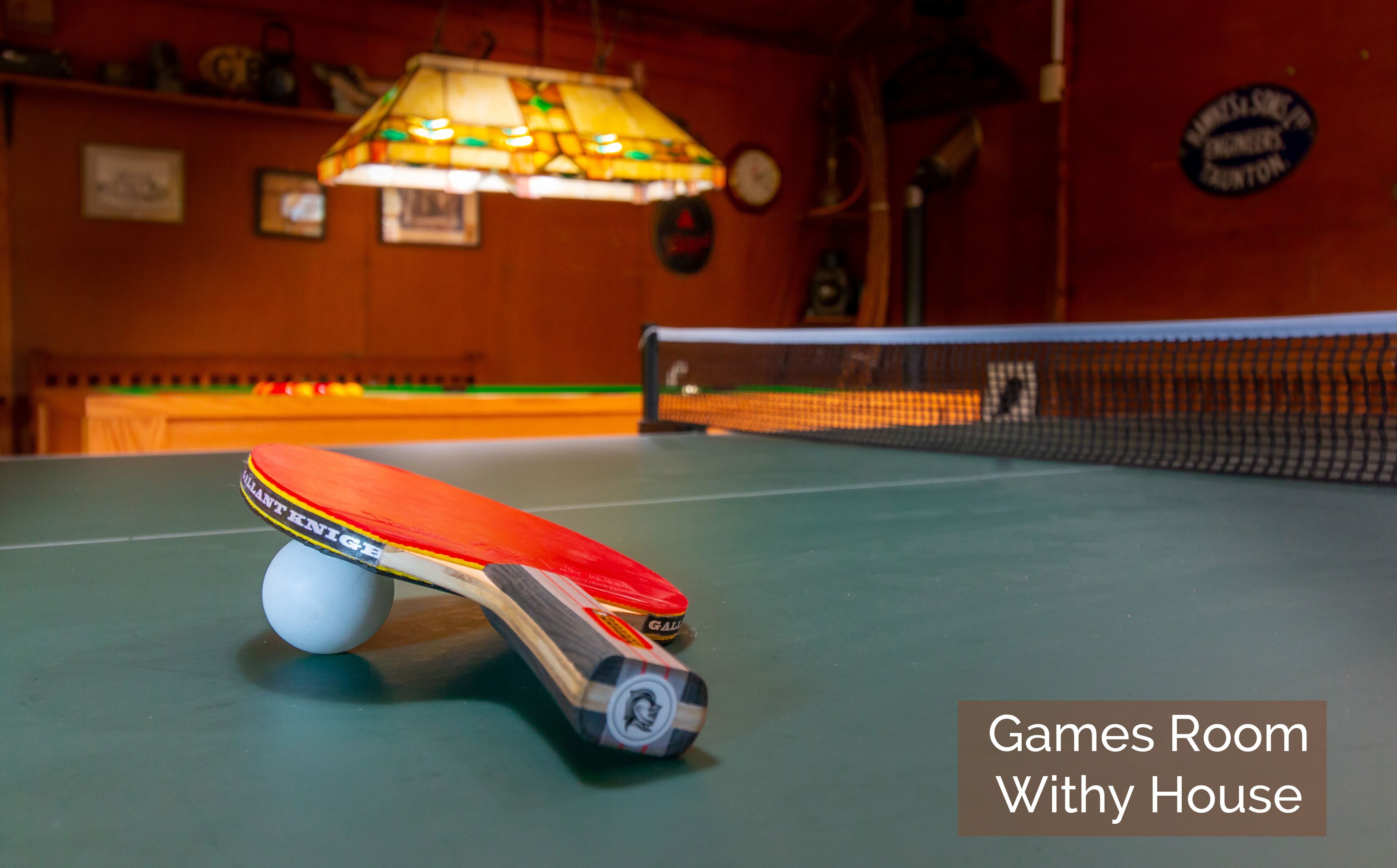 A table tennis table with a paddle and ball in a room with a net. The room has a sign that reads "Games Room Wighty House".