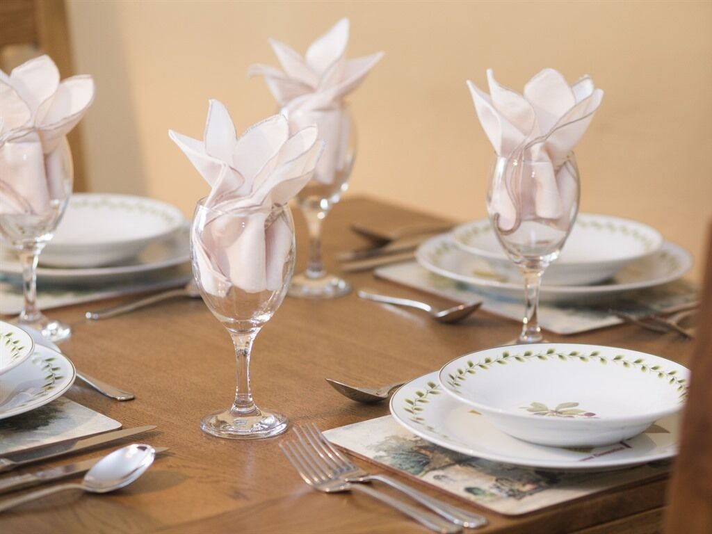 Una mesa de comedor puesta con platos blancos, cristalería transparente y servilletas dobladas en forma de flores.