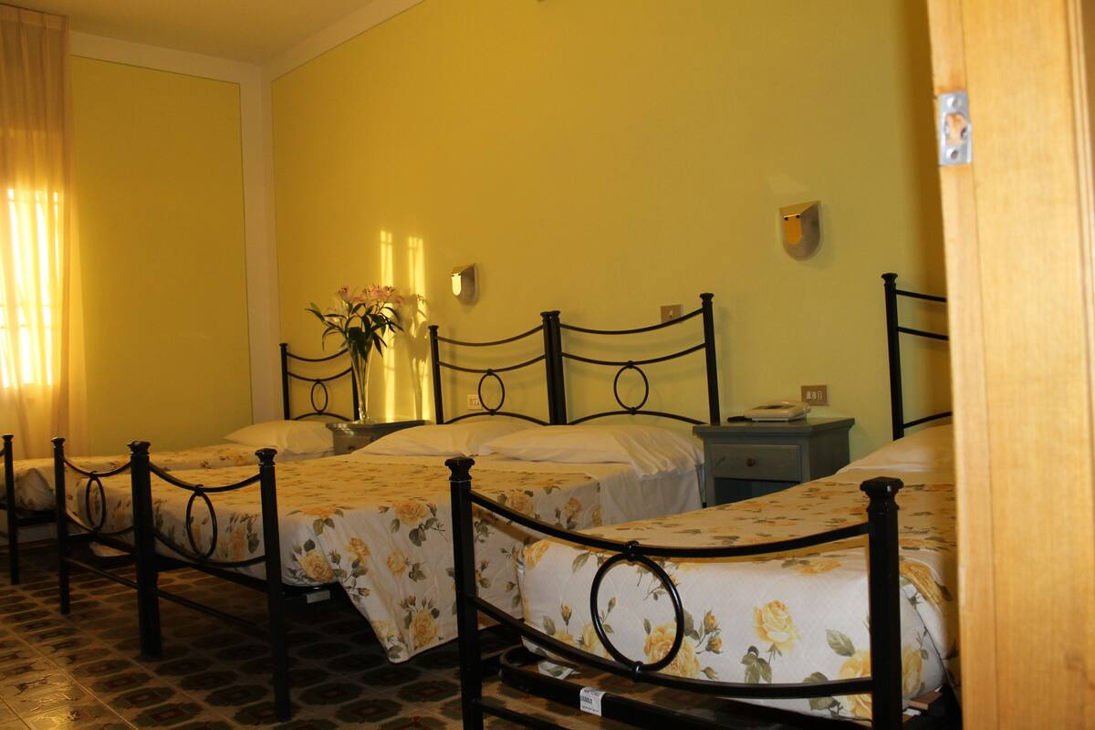 A room with three single beds, each with a floral bedspread and black metal headboards. There is a nightstand with a lamp and a small table with a book.