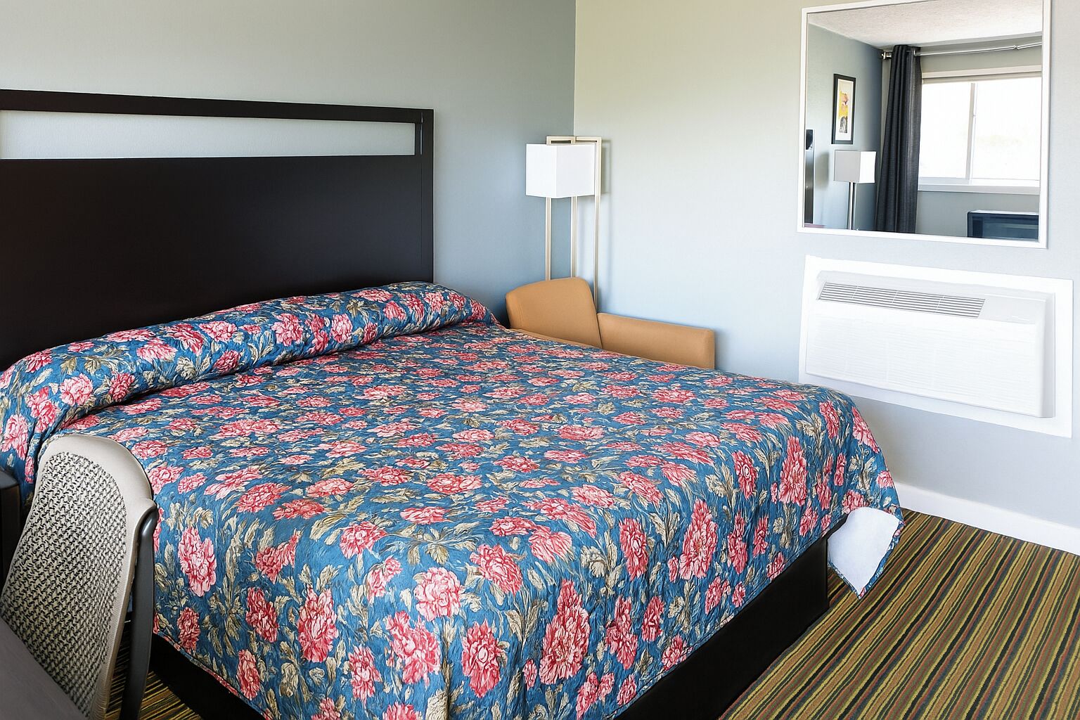 Una habitación de hotel con una cama grande, un cabecero, una mesita de noche, una lámpara, un espejo y un equipo de aire acondicionado.