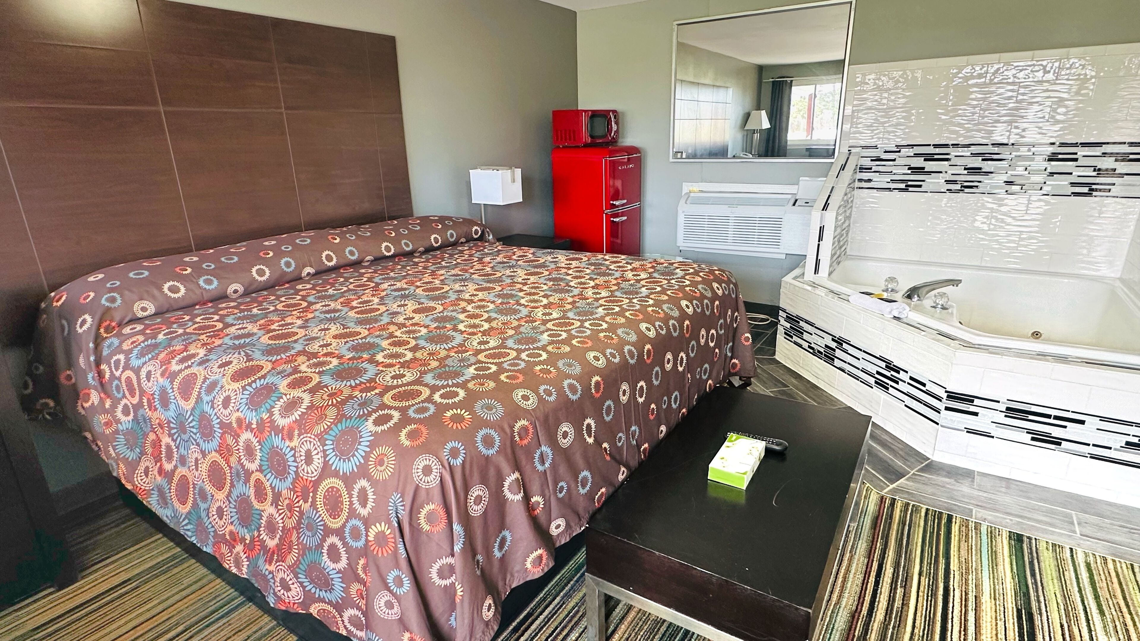 A hotel room with a bed, a red minibar, a white bathtub, and a striped rug.