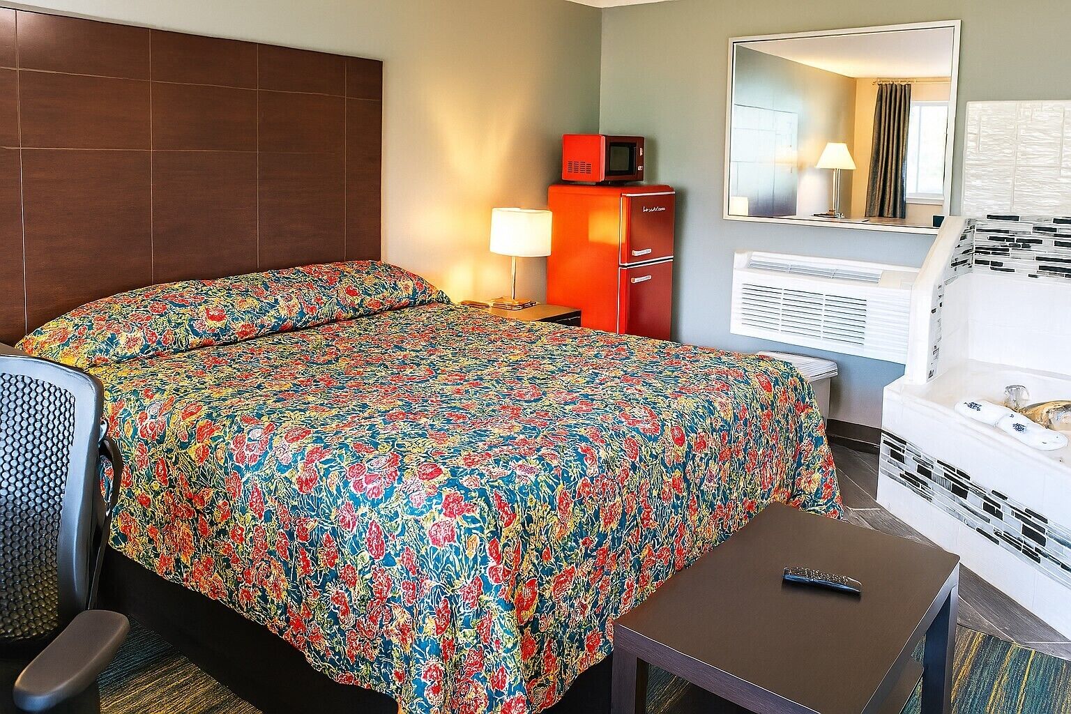 A hotel room with a bed, a small table, a sofa, a microwave, a cabinet, a mirror, and a bathtub.