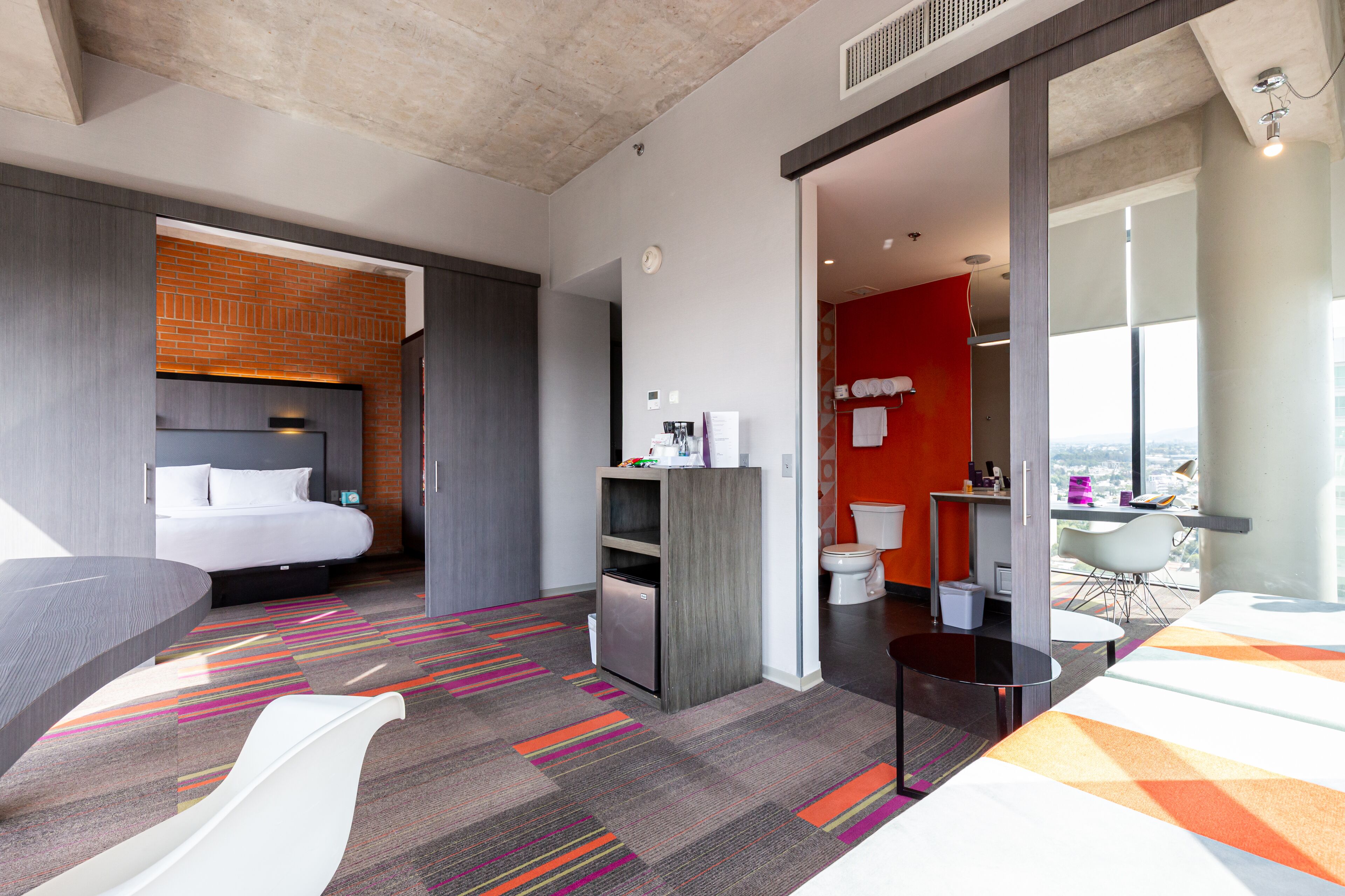 A modern hotel room with a bed, a desk, a bathroom, and a view of the city.