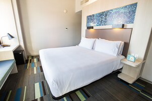 Habitación de hotel con una cama grande, un escritorio con lámpara, una mesita y una obra de arte colgada en la pared.