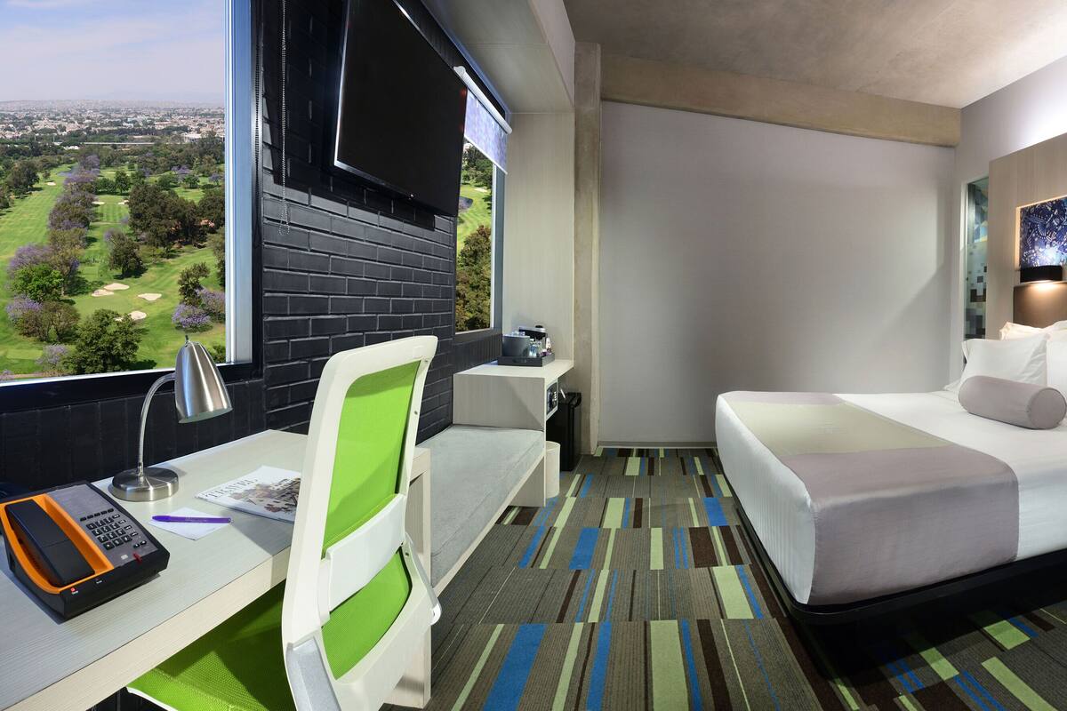 Habitación de hotel con cama, escritorio, silla y un amplio ventanal que ofrece vistas a un campo de golf.
