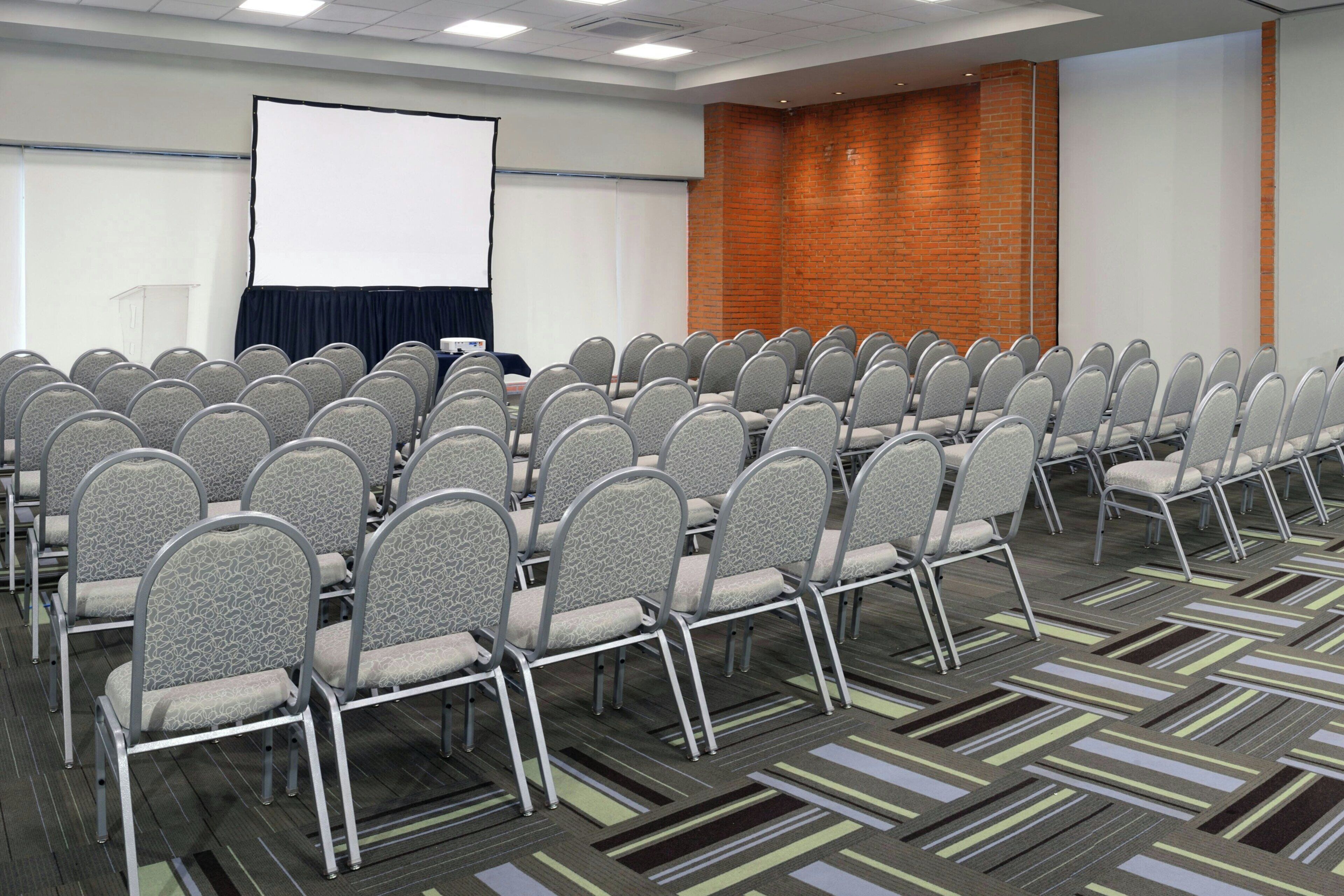 Sala de conferências com fileiras de cadeiras, uma tela de projetor e uma parede de tijolos.