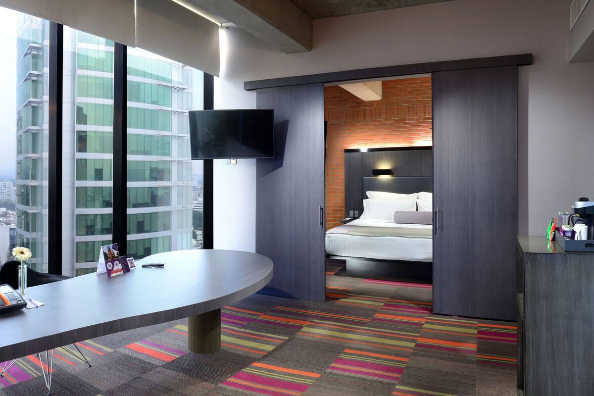 Habitación de hotel moderna con un escritorio grande, una cama, un televisor y vistas de la ciudad a través de amplias ventanas.