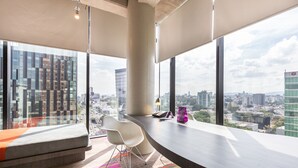 A modern office space with a curved desk, a cushioned bench, and a view of a cityscape through large windows. - Hotel Guadalajara Country Club by HNF (Guadalajara)