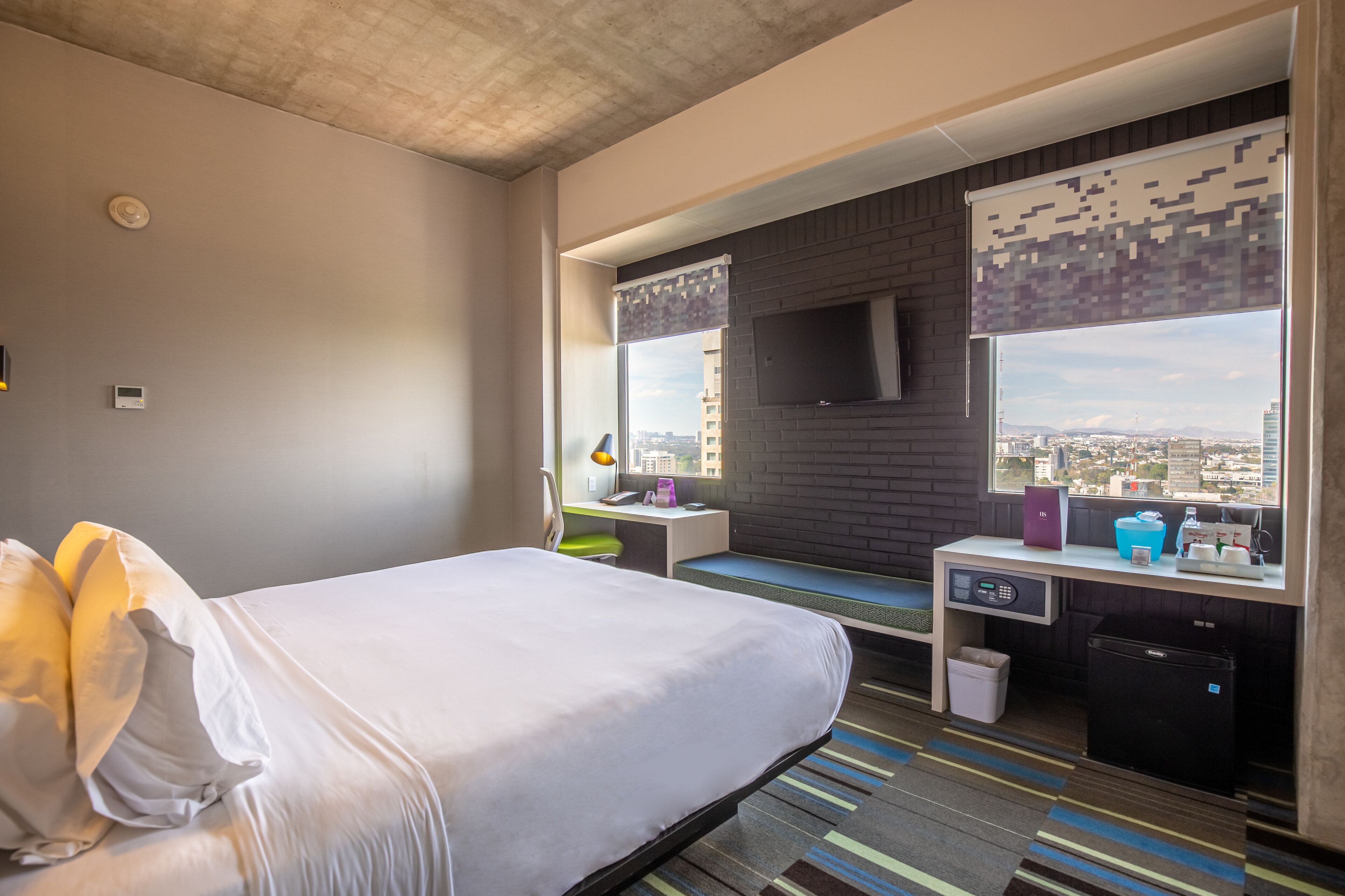 A modern hotel room with a large bed, a desk with a computer, a small fridge, and a window with a city view.