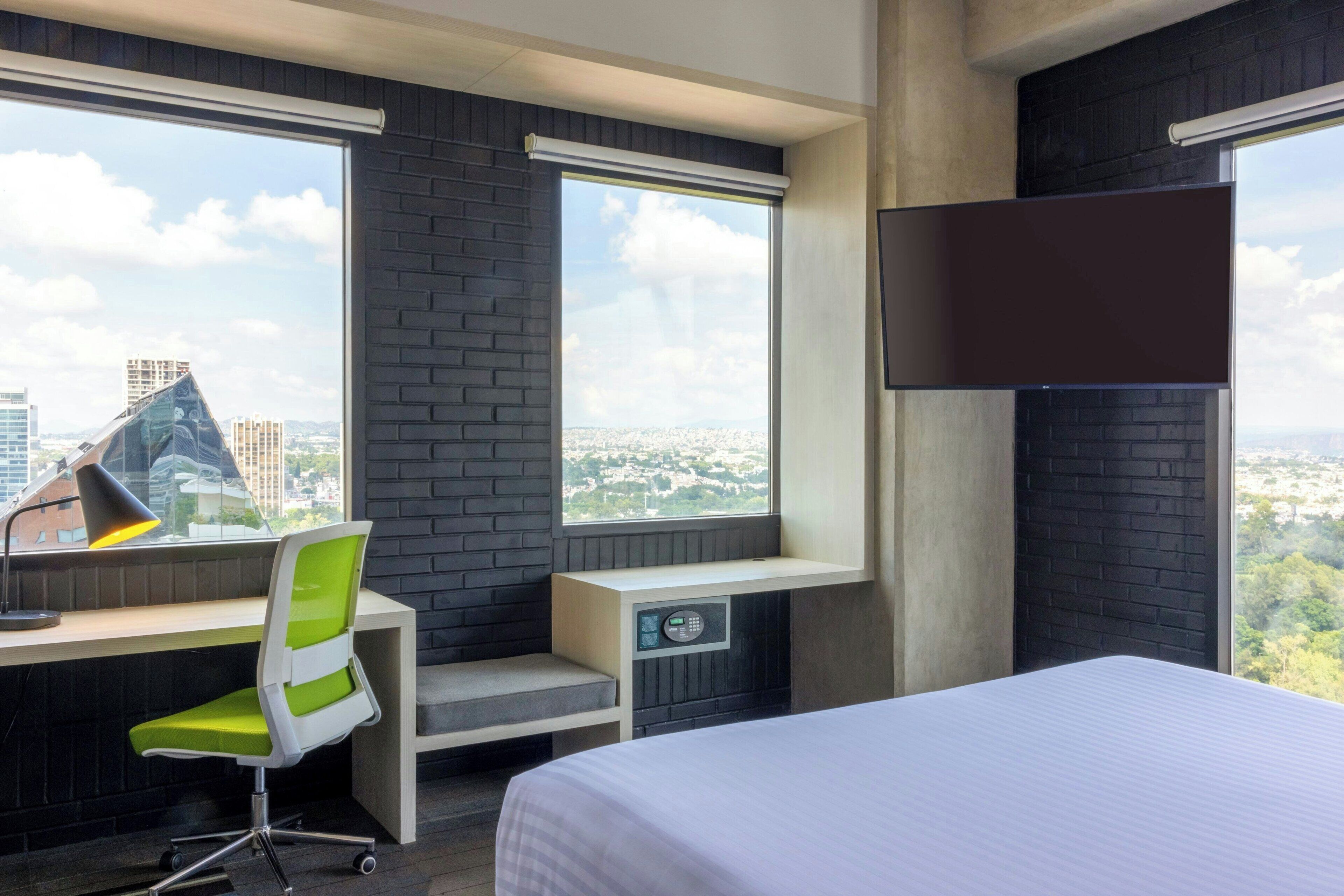 Une chambre d’hôtel dotée d’une grande fenêtre, d’un bureau avec une chaise, d’un lit et d’un téléviseur fixé au mur.