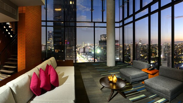 A modern hotel room with a city view, a sofa with pink cushions, a grey armchair, and a small table with decorative vases. - Hotel Guadalajara Country Club by HNF (Guadalajara)