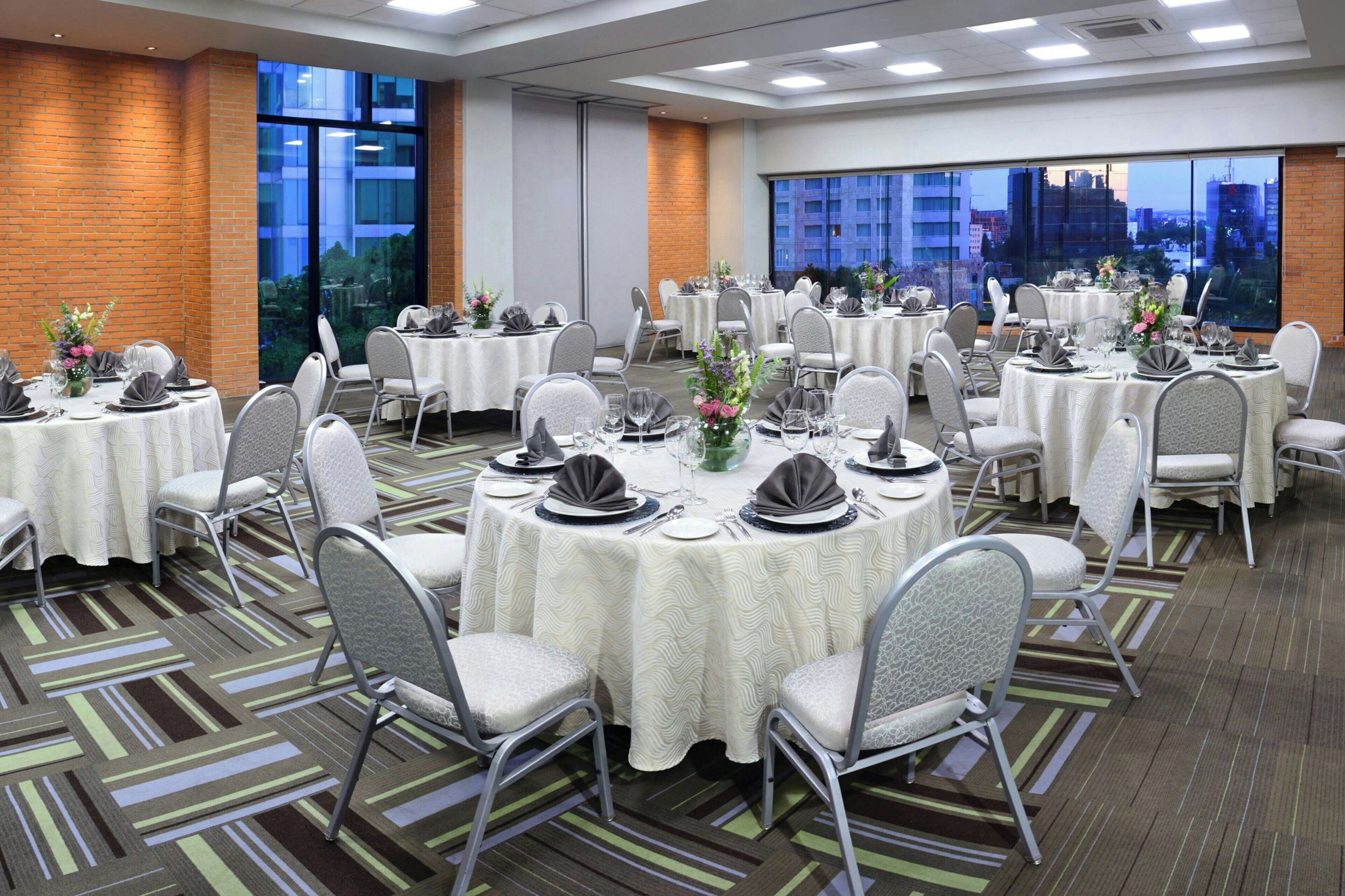 A banquet hall with round tables, chairs, and a view of a cityscape through large windows.