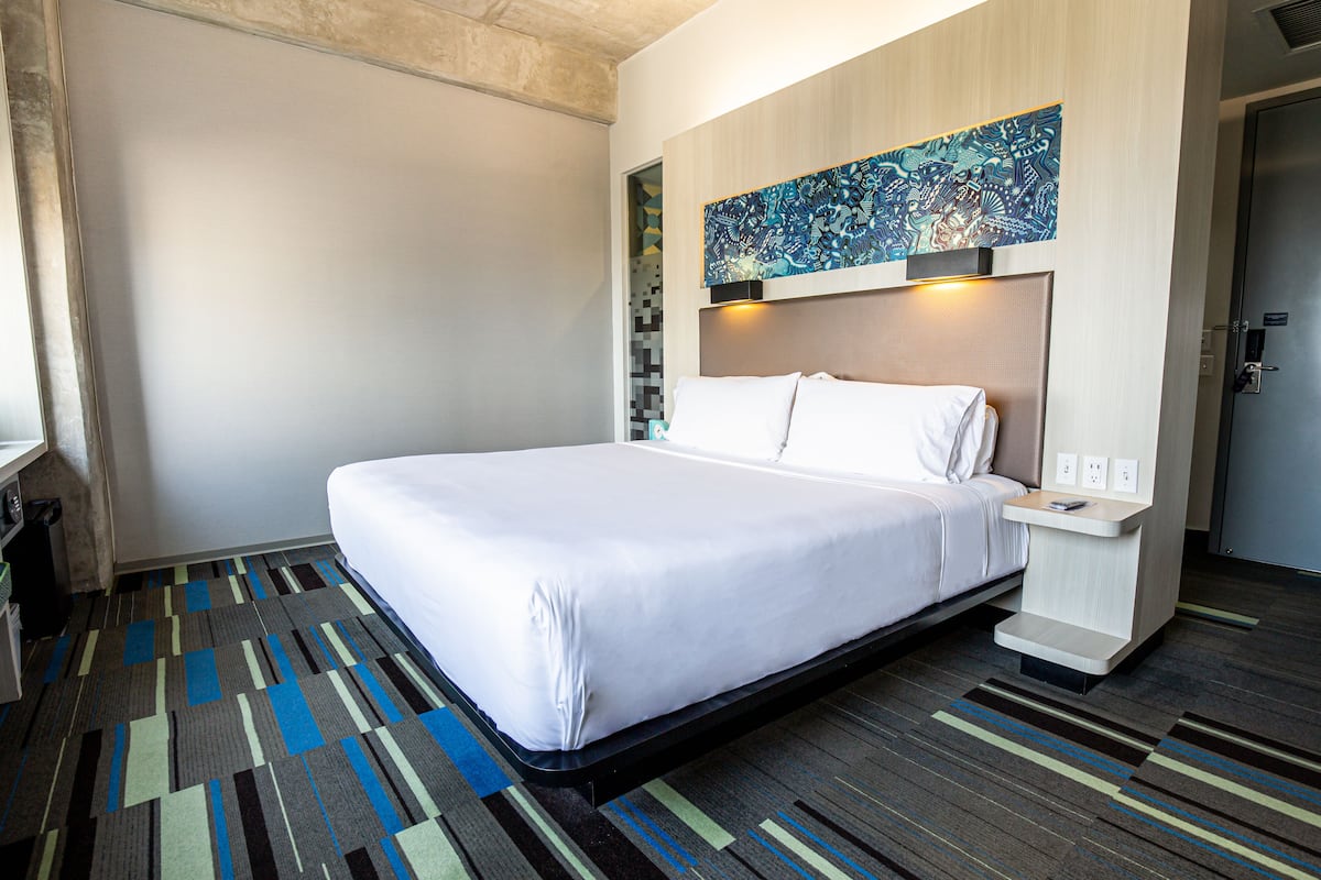 Una habitación de hotel con una cama grande, una mesita de noche, una obra de arte colgada en la pared y una alfombra estampada.