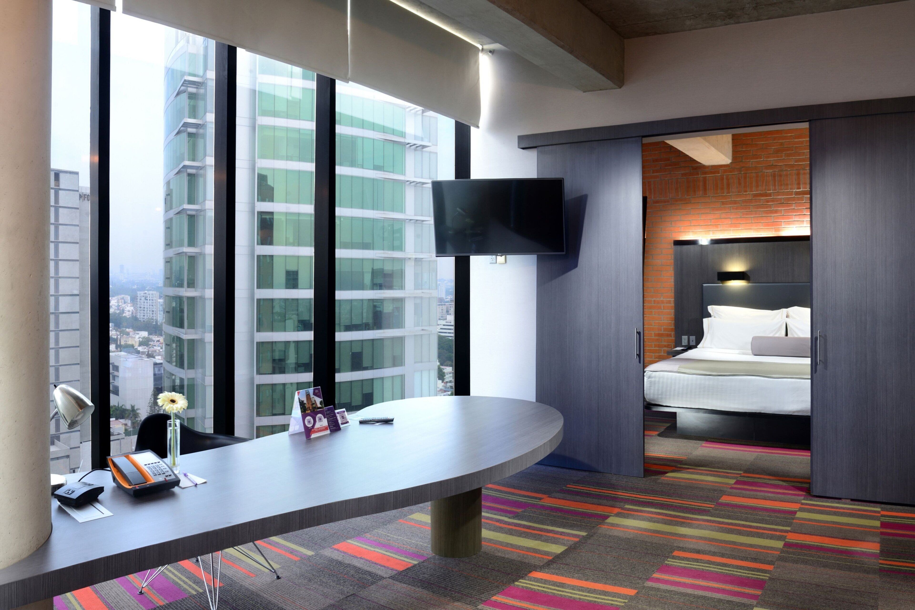 Una habitación de hotel moderna con un escritorio grande, un televisor de pantalla plana montado en la pared, una cama con cabecera y vistas a la ciudad a través de amplias ventanas.