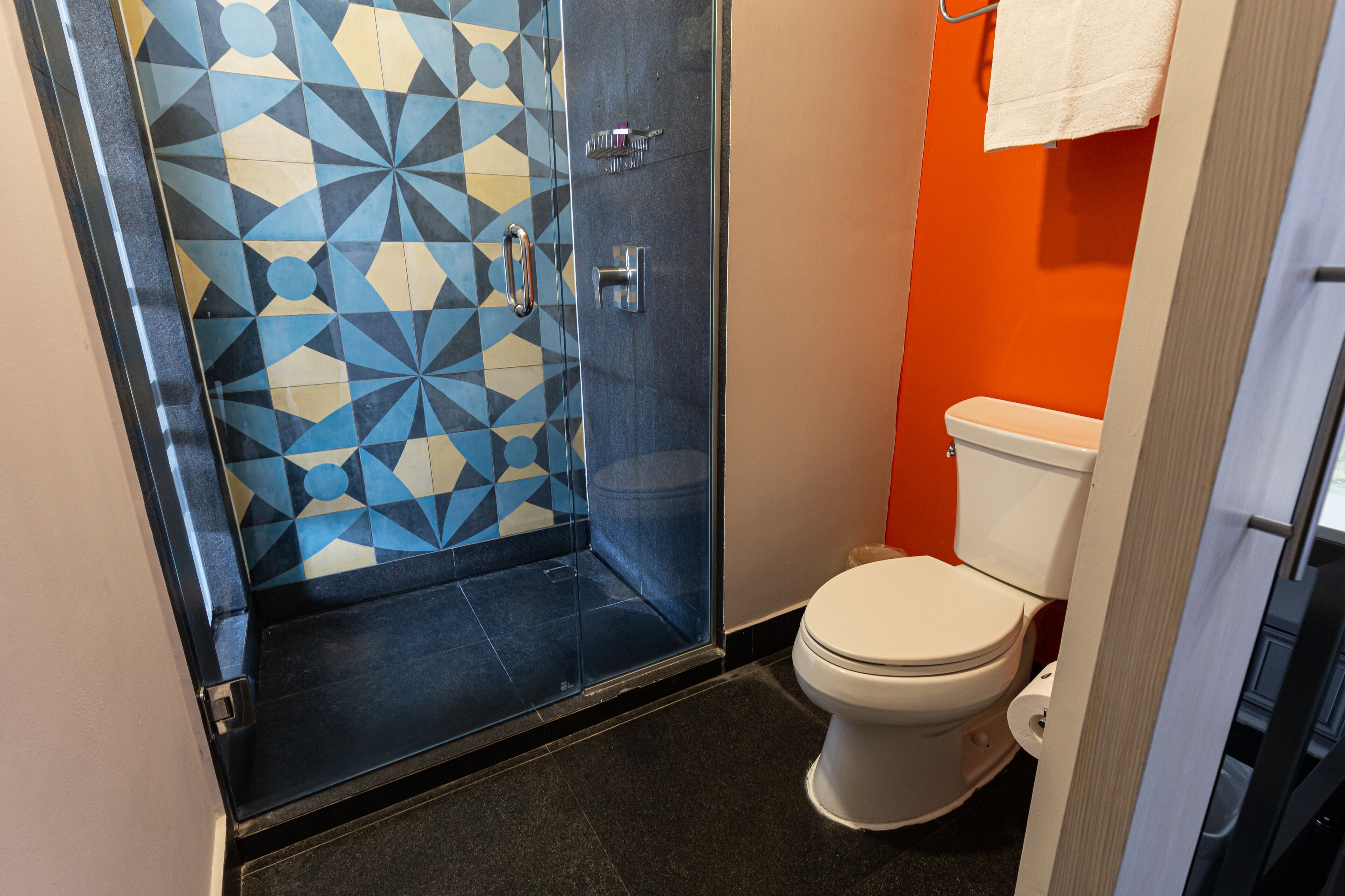 Une salle de bain avec une douche carrelée de motifs géométriques, des toilettes blanches et une serviette accrochée au mur.