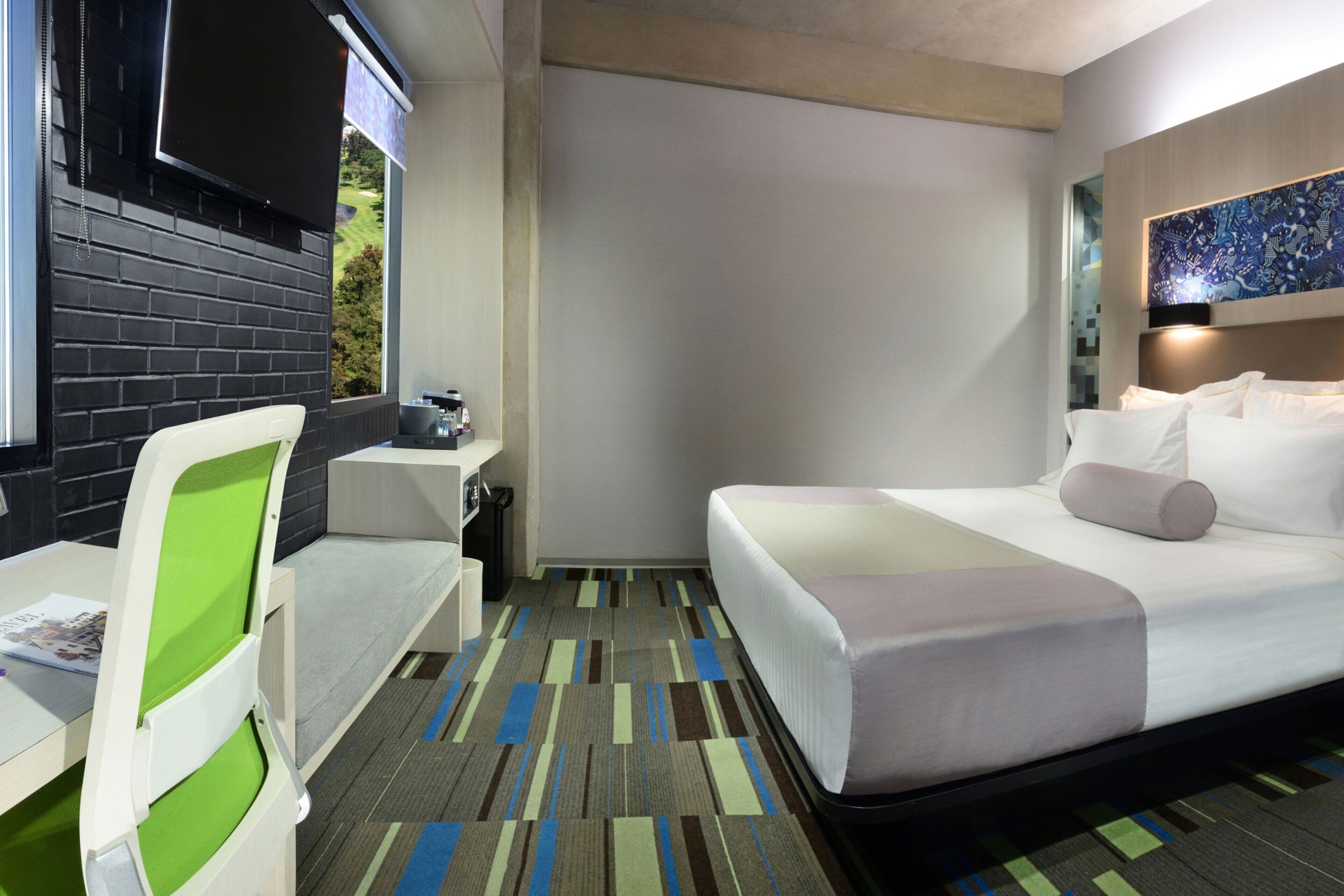 A hotel room with a bed, a desk, a chair, a TV, and a window with a view of greenery.