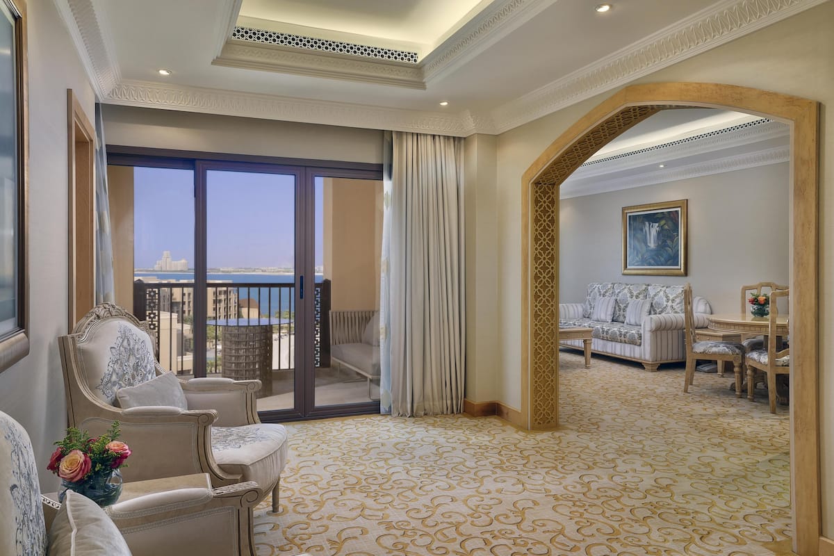 A hotel room with a balcony, a sofa set, a dining table, and a patterned carpet.