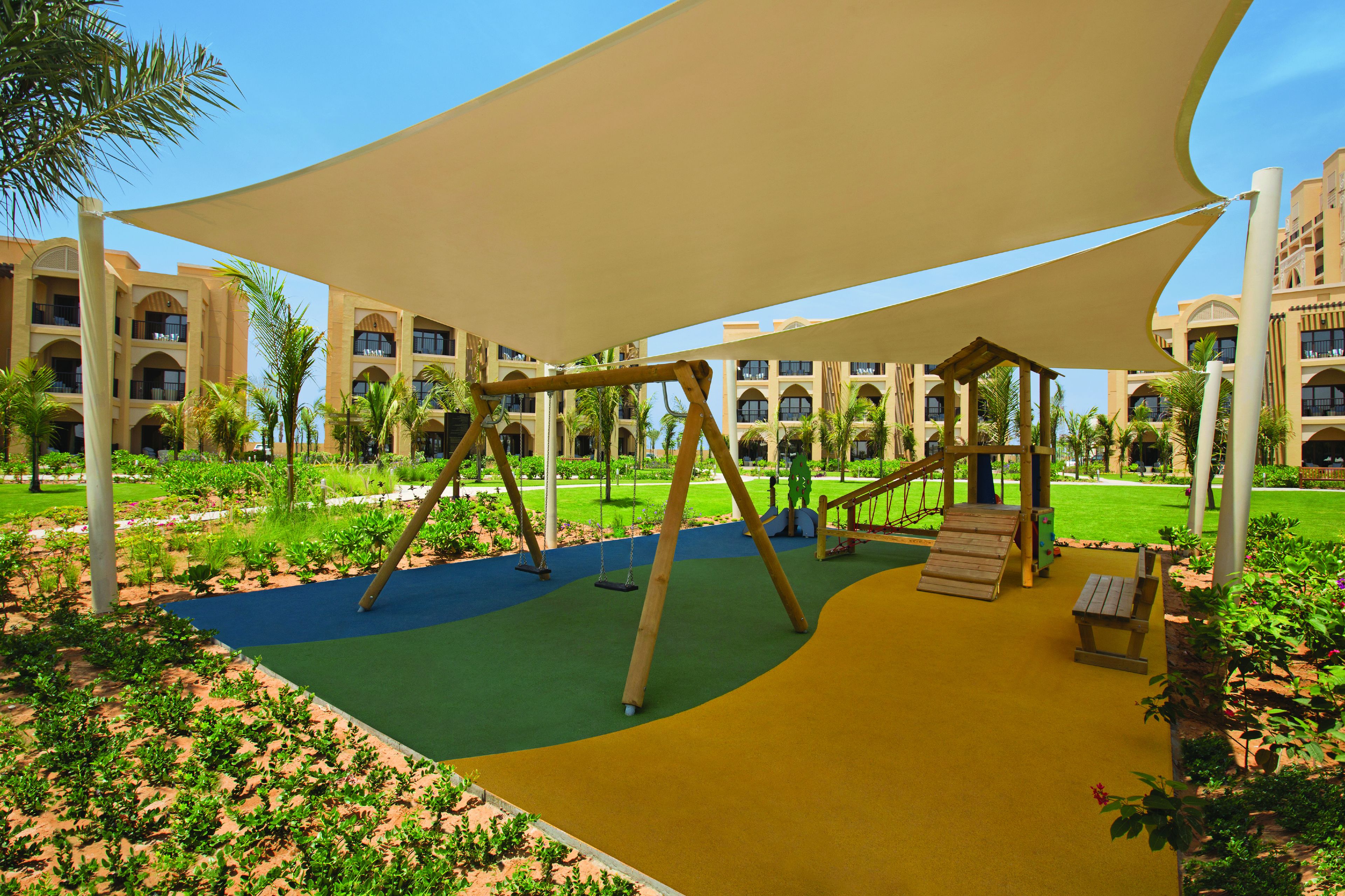 A playground with swings and a slide, surrounded by palm trees and a building with multiple balconies.
