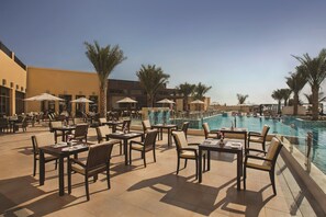 A poolside area with tables and chairs, umbrellas, and a pool.