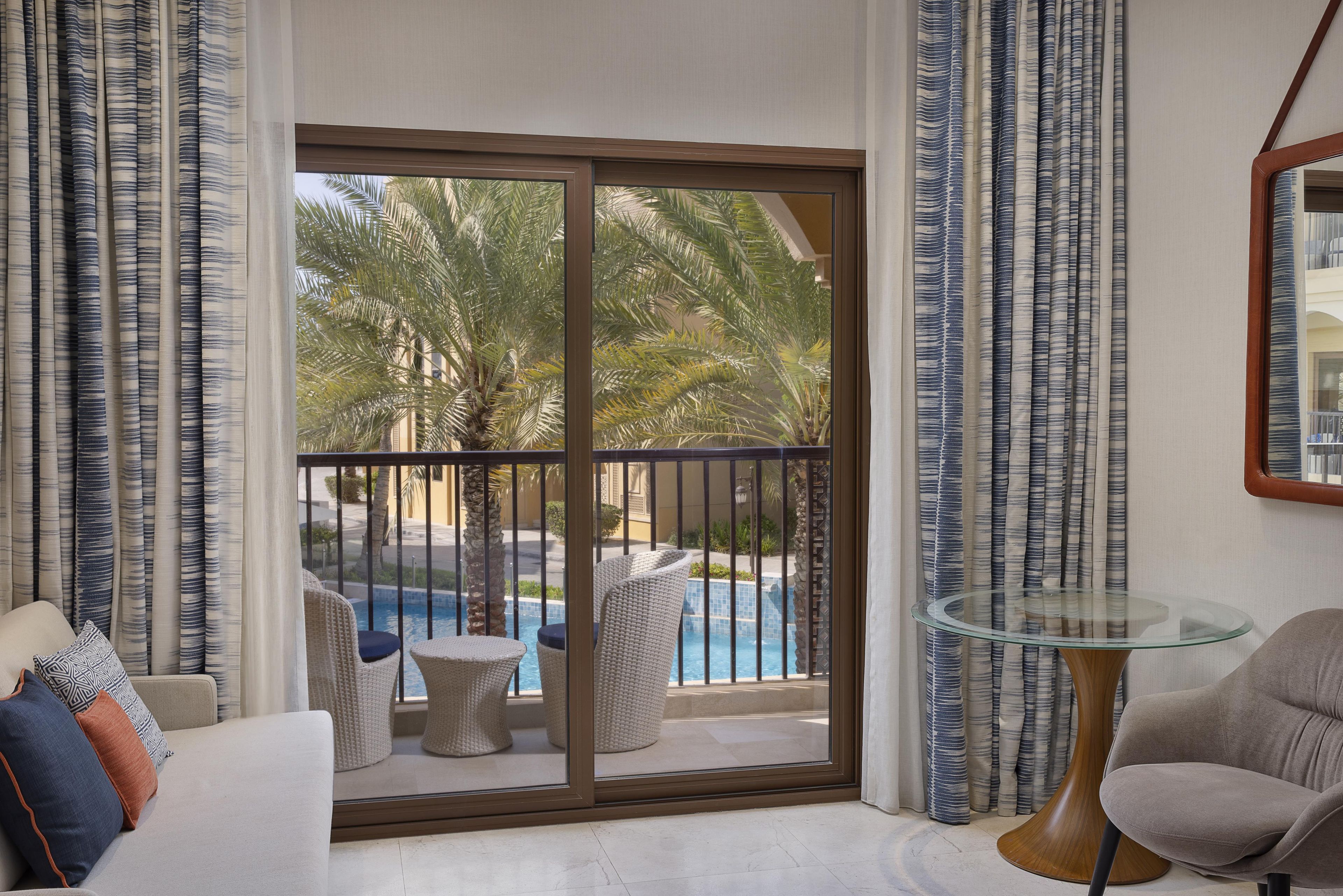 Een hotelkamer met balkon, zwembad, rieten meubels en een glazen tafel.
