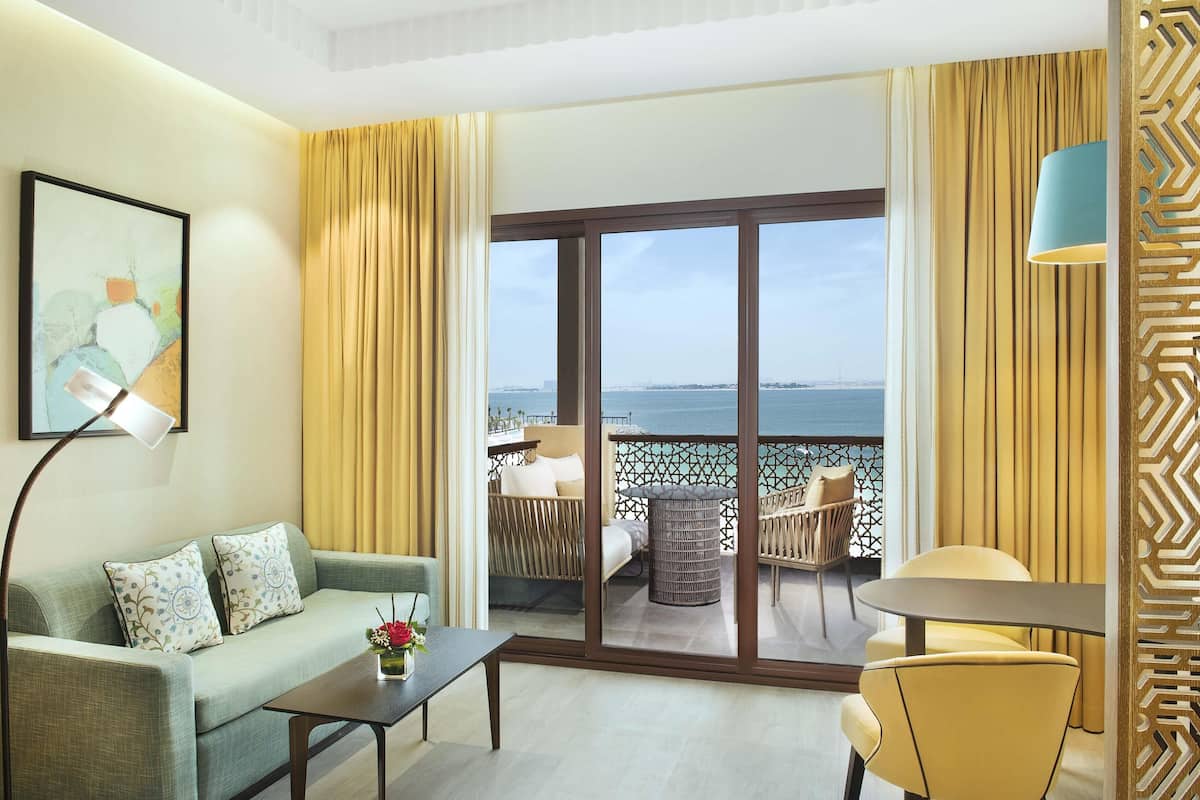 A hotel room with a sofa, a table, a chair, a balcony with a view of the sea, and a large window.