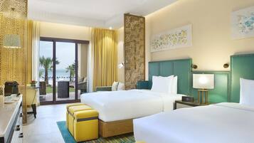 A hotel room with two beds, a desk with a coffee maker, a view of the beach, and a balcony with chairs.