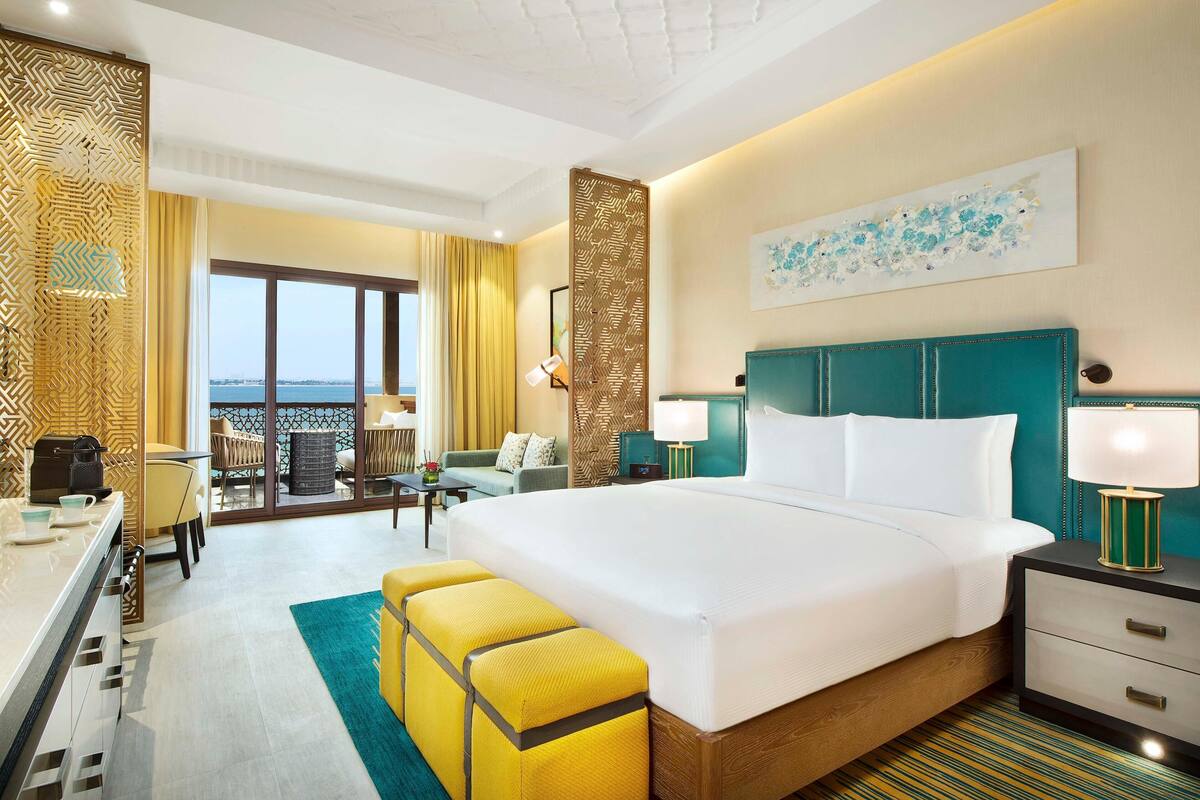 A hotel room with a large bed, a desk with a chair, a balcony with a view, and a dining area with a table and chairs.