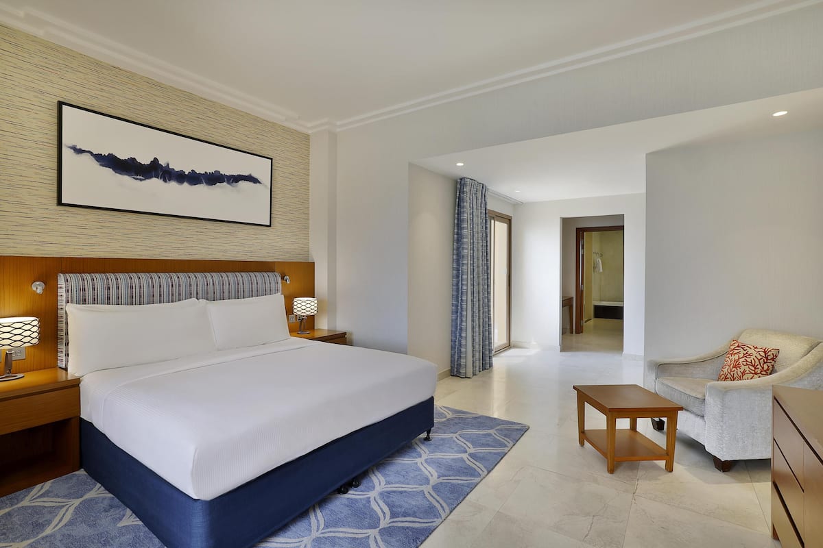 A hotel room with a large bed, a nightstand, a desk, a chair, and a view of the hallway.