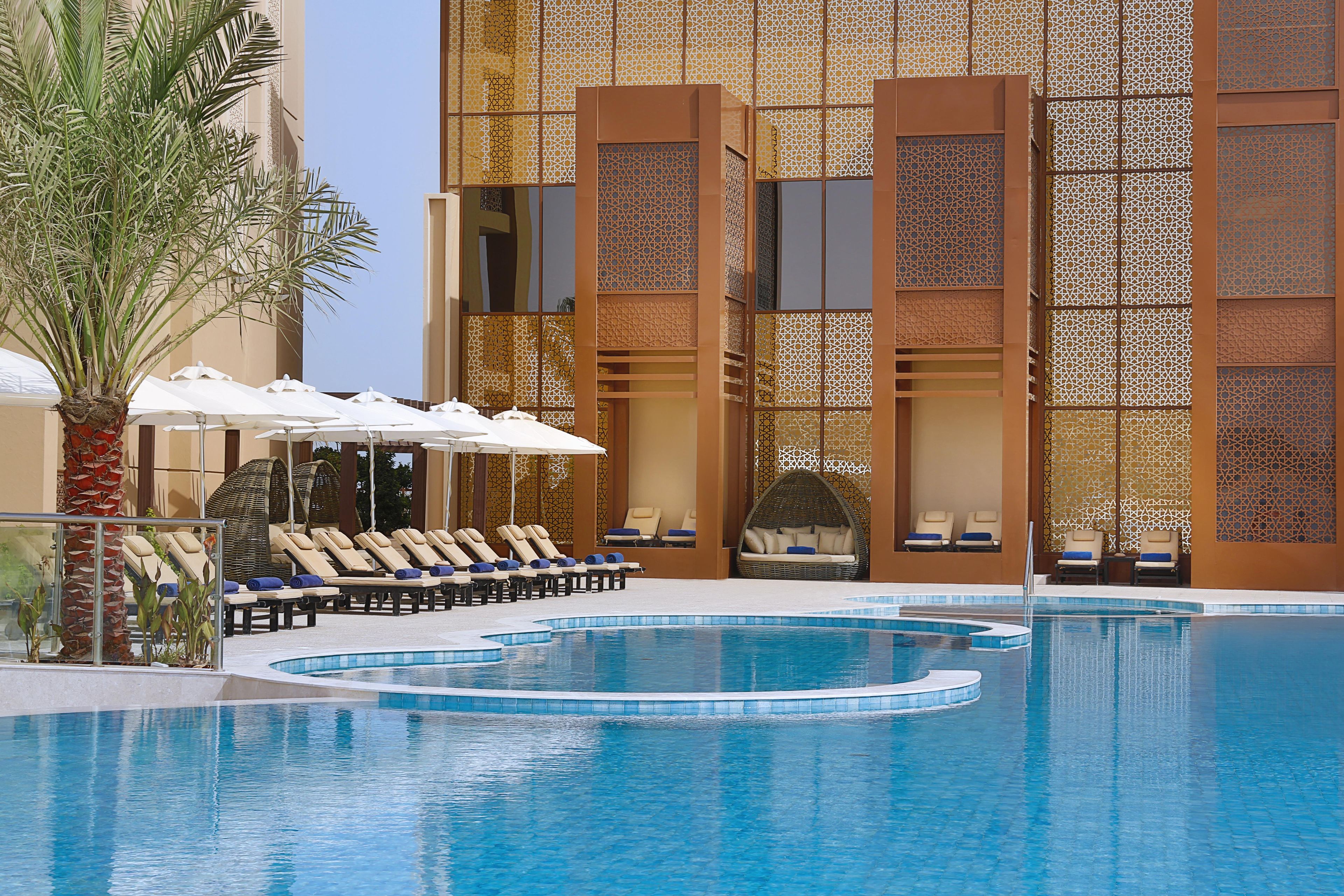 A pool area with lounge chairs and umbrellas, a palm tree, and a building with patterned windows.